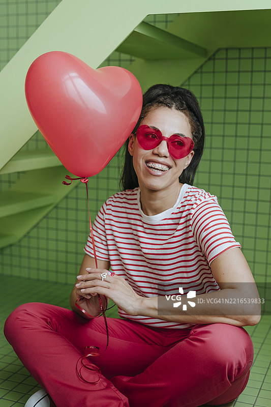 室内微笑的女人，手持心形气球和红色太阳镜图片素材