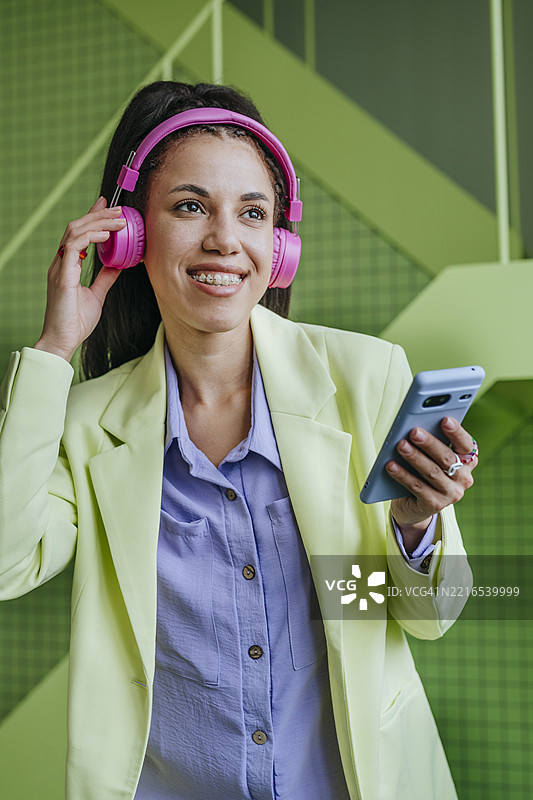 一位女性在室内微笑着，戴着粉色耳机，手里拿着手机听音乐。图片素材
