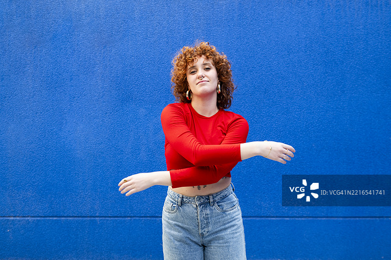 自信的卷发女性穿着红色衬衫，摆姿势于蓝色背景前图片素材