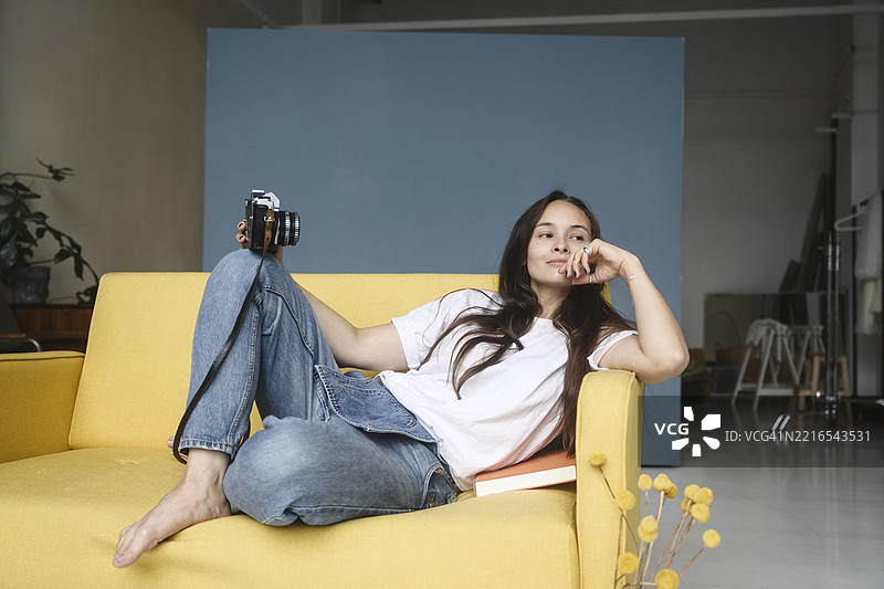 女人坐在室内的黄色沙发上，手里拿着相机。图片素材
