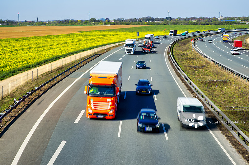 德国于亥根的A44高速公路上，卡车和汽车在运动模糊中穿行，背景是一片油菜花田。图片素材