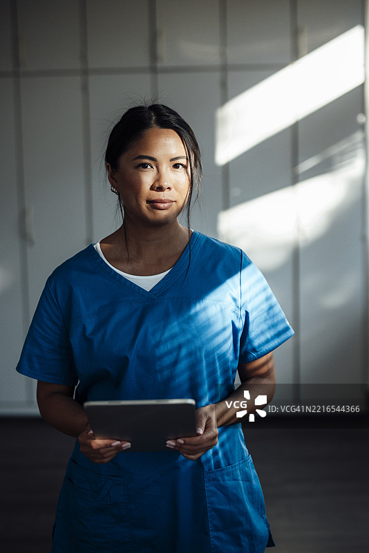 穿着蓝色制服的护士在室内手持平板，阳光透过窗户洒入。图片素材