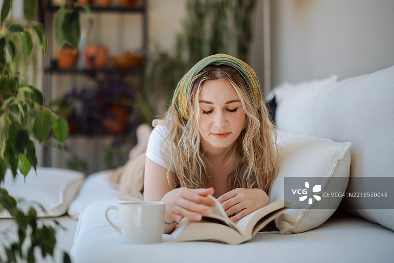 年轻女性躺在沙发上阅读一本书，喝着咖啡图片素材