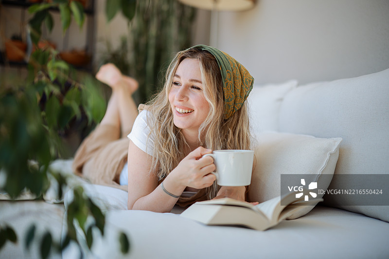 年轻女性躺在沙发上阅读一本书，喝着咖啡图片素材