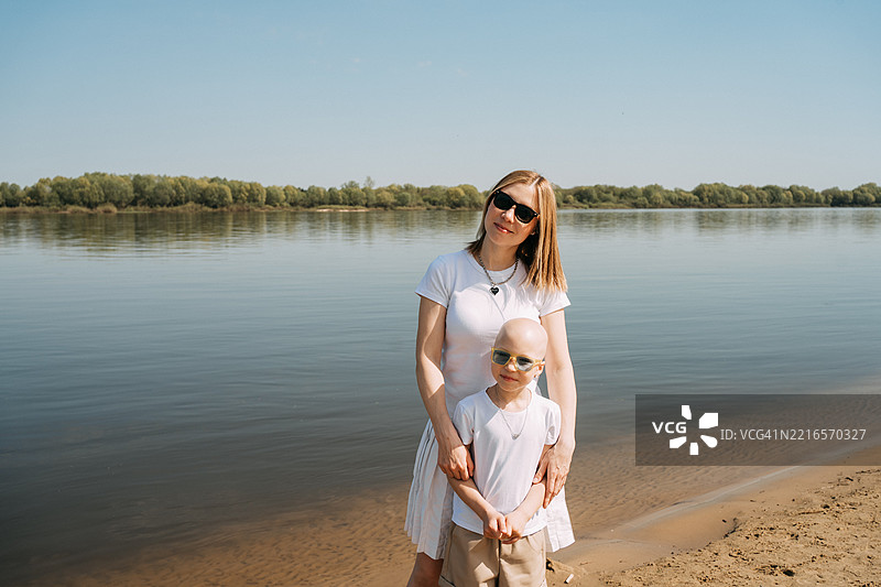 母亲和孩子在河岸上微笑，戴着太阳镜，孩子有脱发症。图片素材