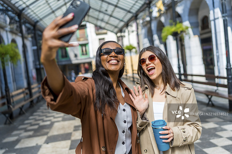 两位快乐的女商人在城市中走路时用智能手机自拍并向镜头挥手。图片素材