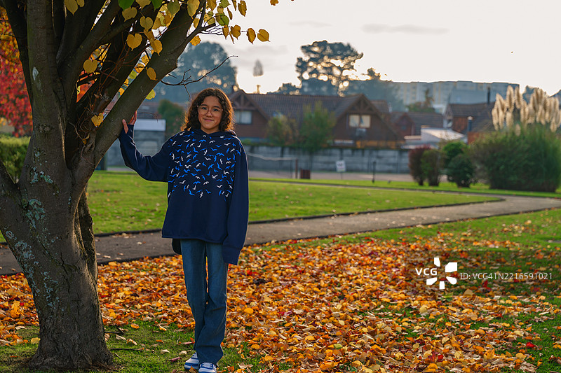 年轻女孩在秋天的公园里靠着树，夕阳西下。图片素材