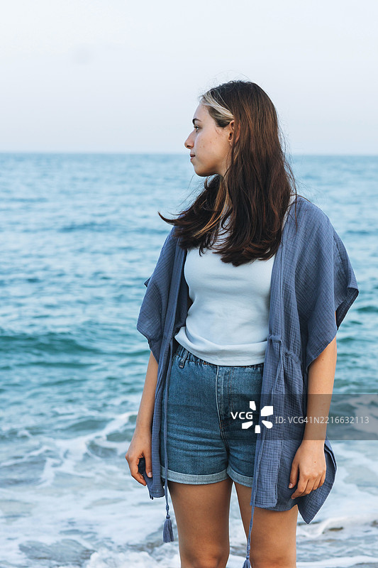 一位穿着休闲服的女性站在海边，若有所思地凝视着海面。图片素材