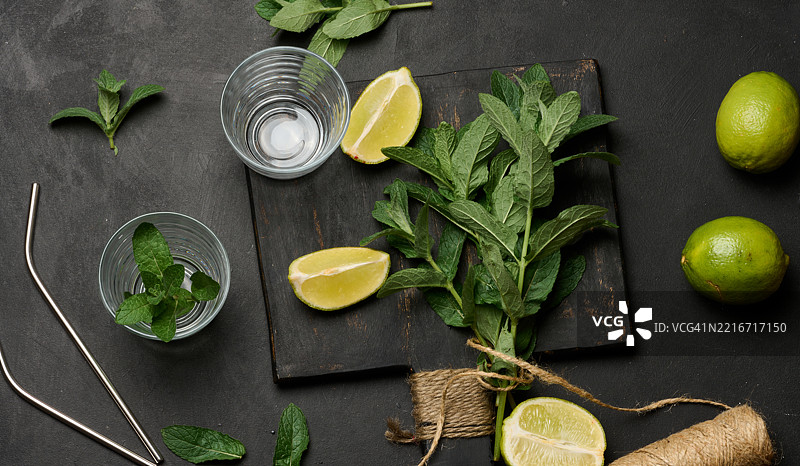 新鲜的绿薄荷束放在木板上，旁边是青柠角、玻璃杯和金属吸管，俯视图的鸡尾酒配料图片素材