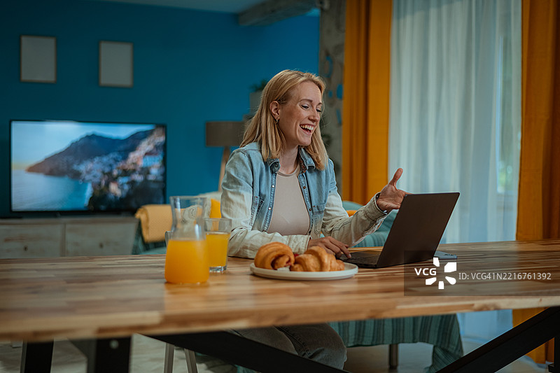 快乐的女人在客厅里边吃早餐边使用笔记本电脑图片素材