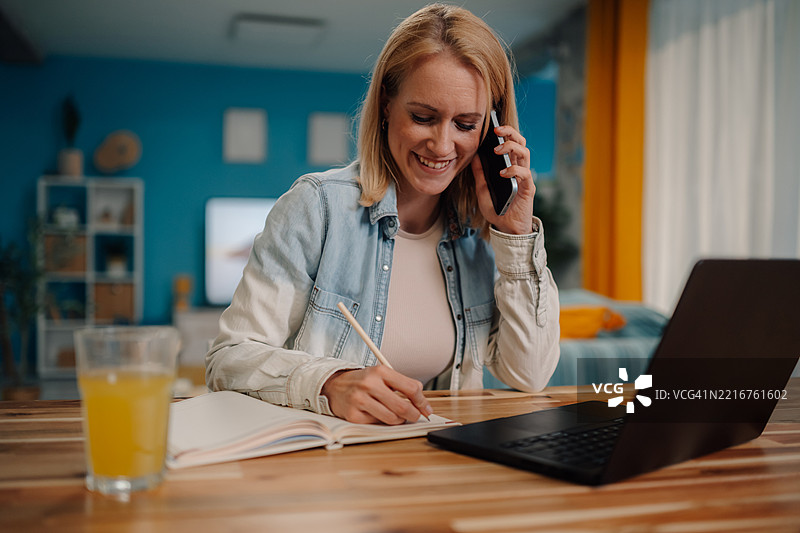 微笑的女性在家中办公室一边打电话一边做笔记图片素材