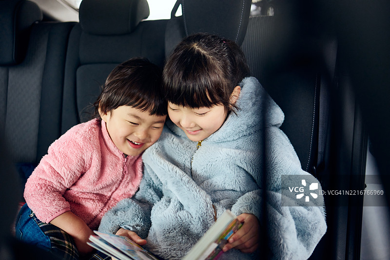 两个年轻的兄妹紧紧坐在汽车后座，头碰着头，充满好奇和快乐地分享一本书。图片素材