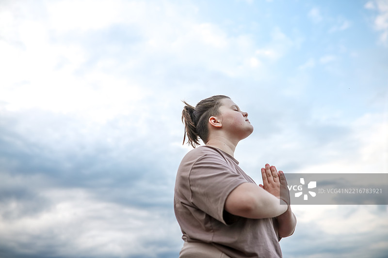 闭眼的胖女孩在天空下放松，双手交叉在胸前。图片素材