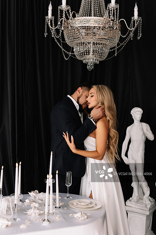 婚礼装饰创意。一对新郎新娘身着优雅的婚礼礼服，在装饰精美的桌子旁相拥亲吻，背景是深色的布料和吊灯。一个男人和一个女人在婚礼仪式上相爱。图片素材