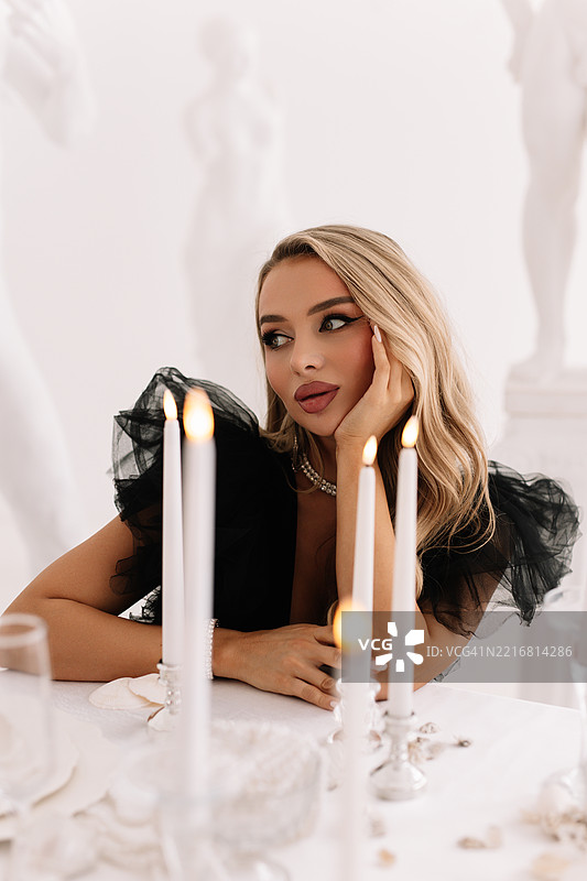 一位美丽年轻女性的肖像，她梳着发型，化着妆，穿着黑色连衣裙，坐在装饰精美的桌子旁，桌上点着蜡烛。图片素材