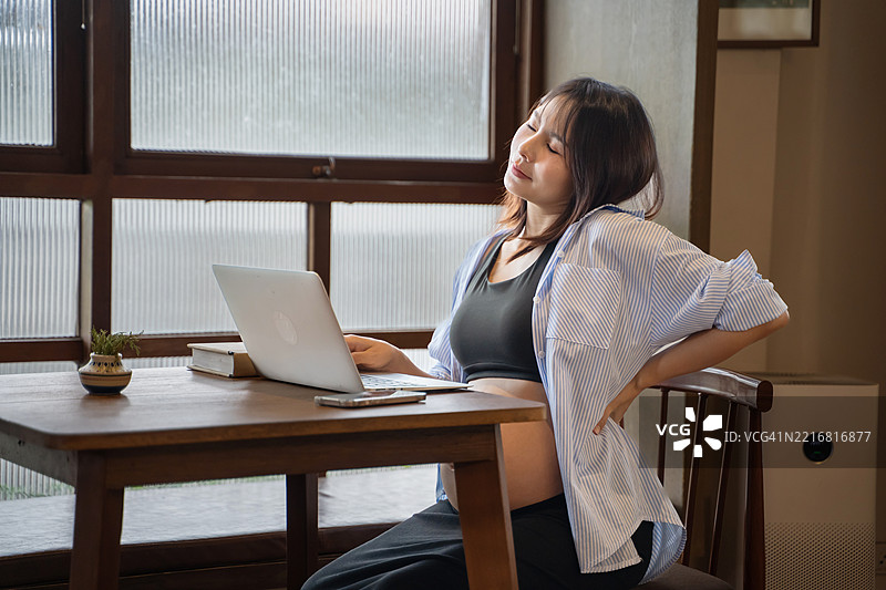 亚洲孕妇因在家办公时坐在桌子前使用电脑而感到背部疼痛。图片素材