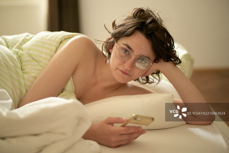 戴眼镜的年轻女性躺在床上，面带平静的表情看着手机。图片素材