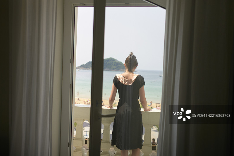 穿着黑色连衣裙的女人站在阳台上俯瞰海滩和海洋图片素材