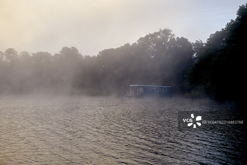 雾气弥漫的湖景，蓝色的房船和周围茂密的树木在柔和朦胧的天空下。梅克伦堡湖区，梅克伦堡-前波美拉尼亚，德国图片素材