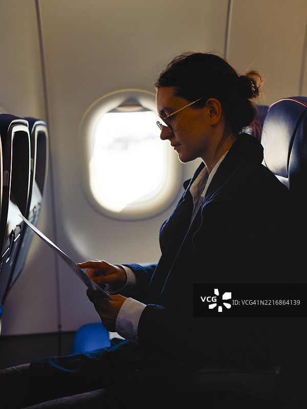 女性在飞机上坐着阅读安全说明，神情平静专注，灯光昏暗。图片素材