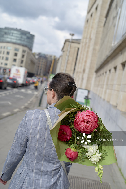 女人在城市街道上走，手里拿着一束绿色包装的粉色花束图片素材