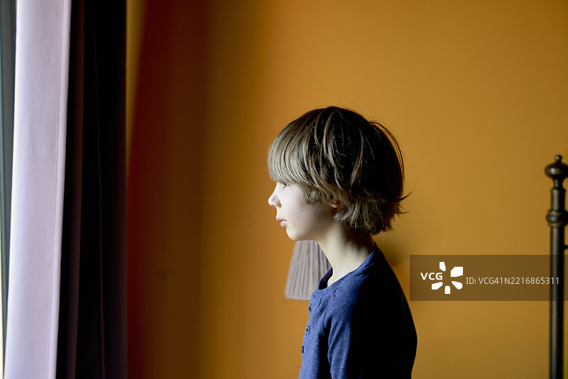 年轻男孩凝视窗外，背景是一面生动的橙色墙壁，房间宁静。意大利西西里。图片素材