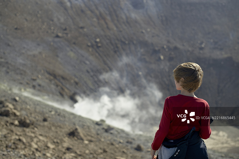 穿着红色衬衫的孩子站在冒烟的火山口前凝视。意大利西西里岛图片素材
