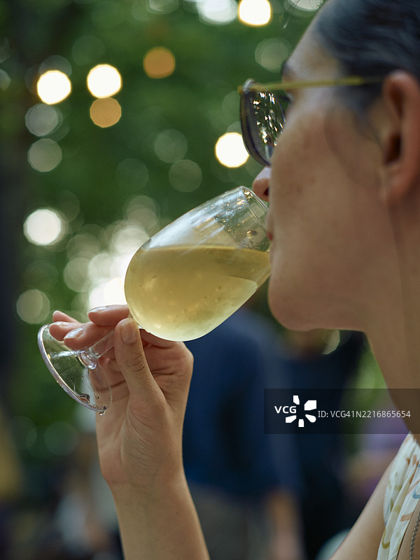 一个戴着太阳镜的人在户外喝着白葡萄酒，背景是模糊的绿树。图片素材