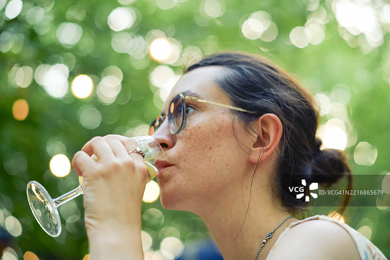 戴眼镜的女人在花园中品尝白葡萄酒，背景模糊。图片素材