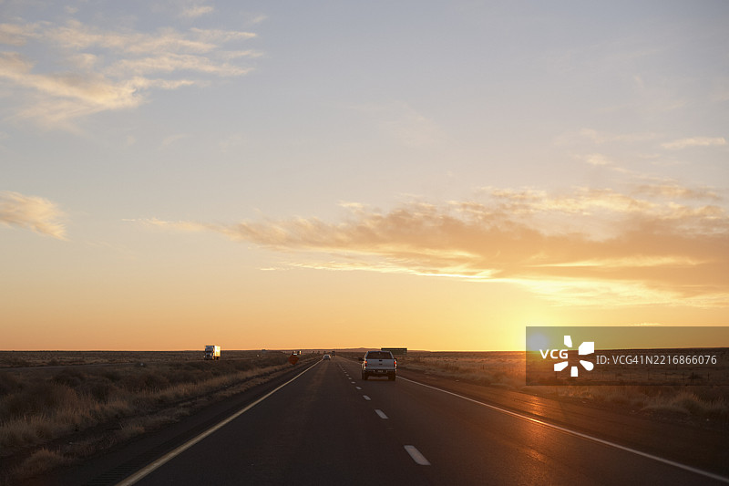 日落时分，开放公路上汽车在金色天空和零星云朵下行驶。美国公路旅行图片素材