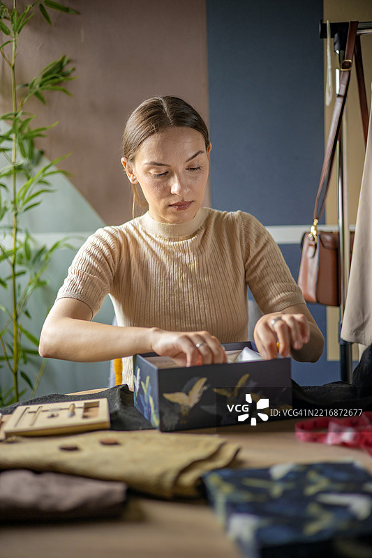 一位年轻的女商人在她舒适的工作室里小心翼翼地将定制服装打包进盒子，准备发货，作为她不断发展的副业的一部分。图片素材