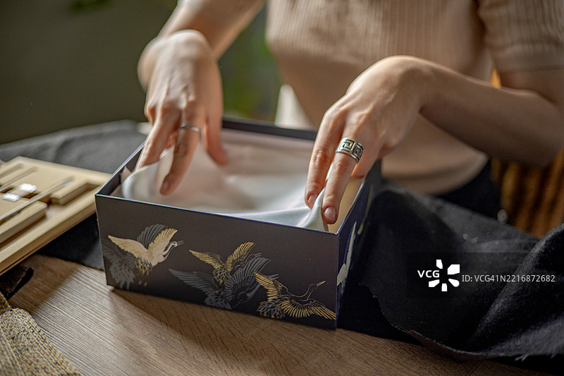 在她温馨的工作室里，年轻女性小心翼翼地将一件完成的衣物装入盒子，准备寄送给她的客户。图片素材