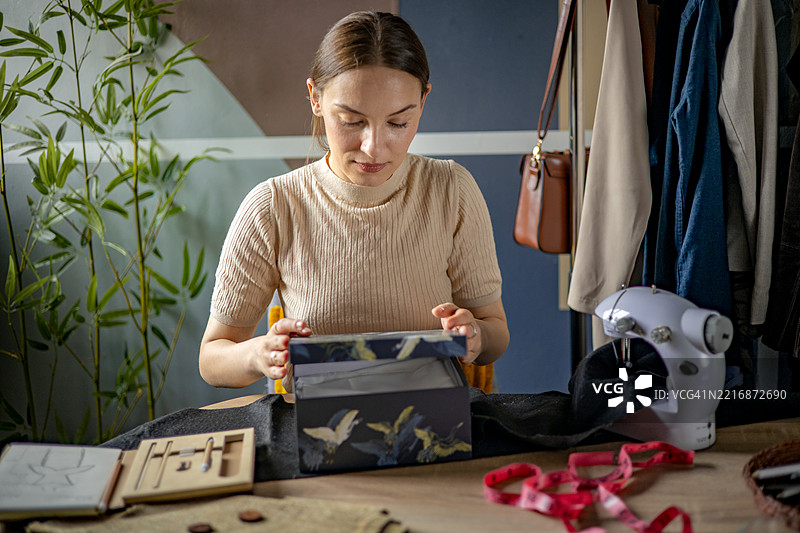 年轻女子被缝纫工具包围，小心翼翼地将完成的衣物包裹起来，放入盒子中，为她的副业准备发货。图片素材