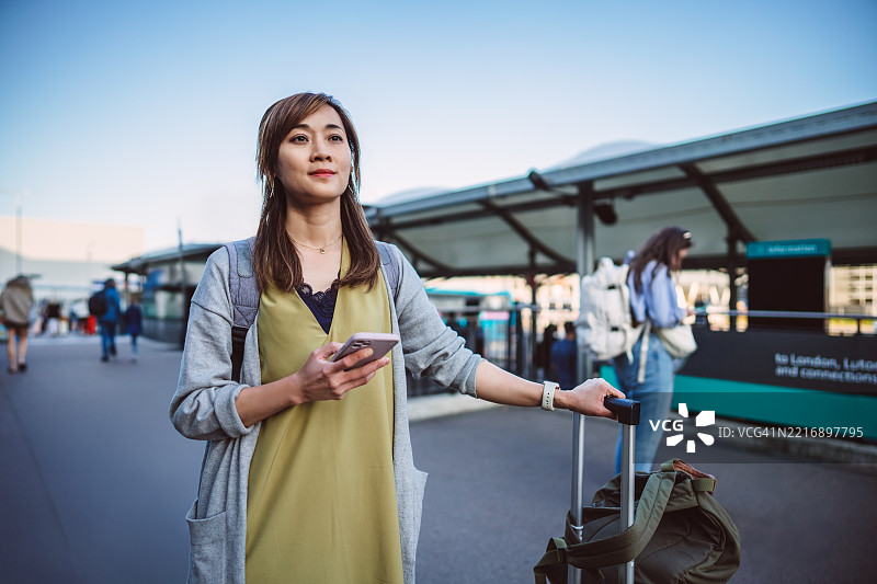 一位女性在交通站独自旅行，手持智能手机和行李。图片素材