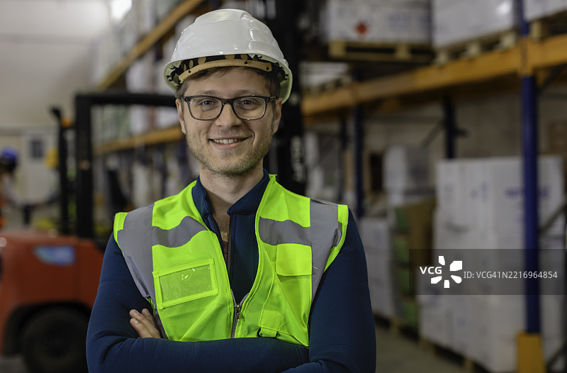 自信的年轻男性仓库工人穿着安全背心和安全帽，双臂交叉在物流中心摆姿势。图片素材
