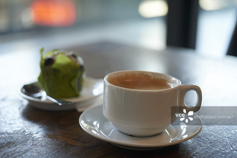 白色咖啡杯静静地放在深色木质咖啡桌上，旁边是一个放在碟子上的开心果松饼，营造出温馨的早晨咖啡氛围。图片素材