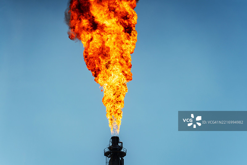 一座巨大的工业厂房被高耸的机械和管道环绕，火焰从燃烧塔喷射而出，照亮了天空。图片素材