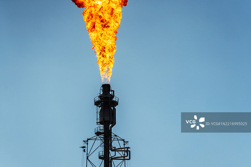 一座巨大的工业厂房被高耸的机械和管道环绕，火焰从燃烧塔喷射而出，照亮了天空。图片素材