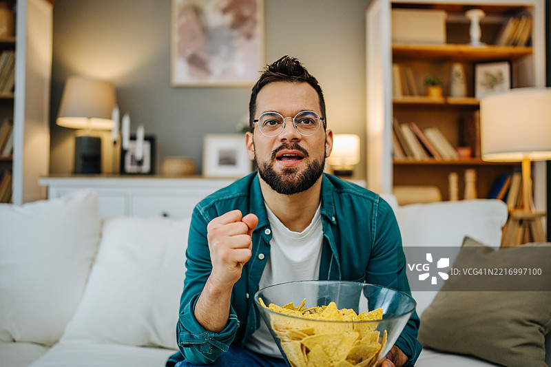 兴奋的男人在家看电视时欢呼并吃着玉米片图片素材