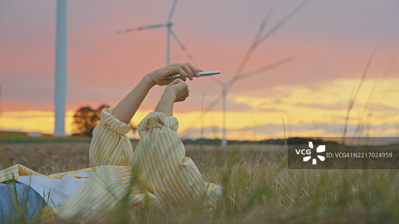 年轻女性在金色日落时分的草地上放松，身旁是风力发电机图片素材