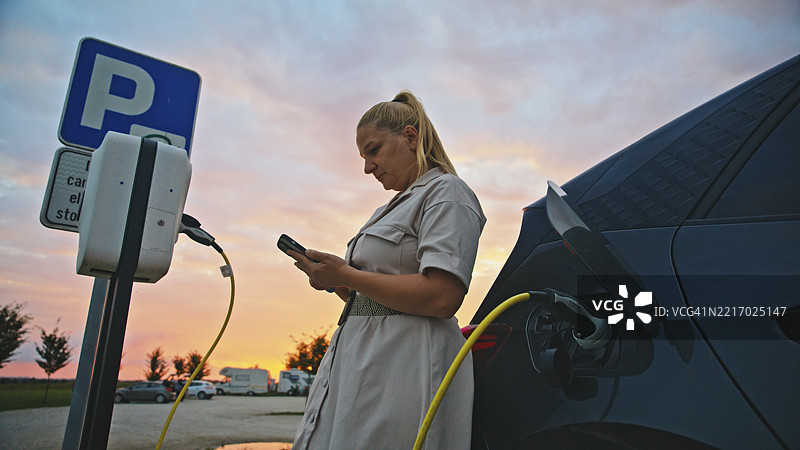 金发女性穿着米色外套，在日落的停车场使用智能手机，同时电动车正在充电。图片素材