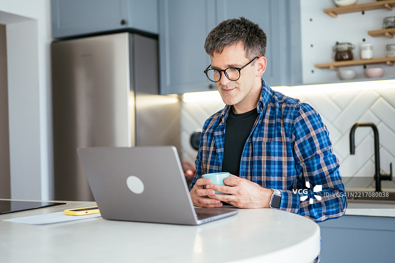 一位穿着蓝色格子衬衫、戴着眼镜的休闲男士在温馨现代的厨房里一边喝咖啡，一边看着笔记本电脑。图片素材