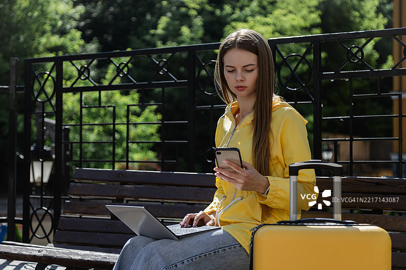 年轻女子穿着黄色雨衣，坐在公园里，手里拿着智能手机，旁边有一个笔记本电脑和一个行李箱。图片素材
