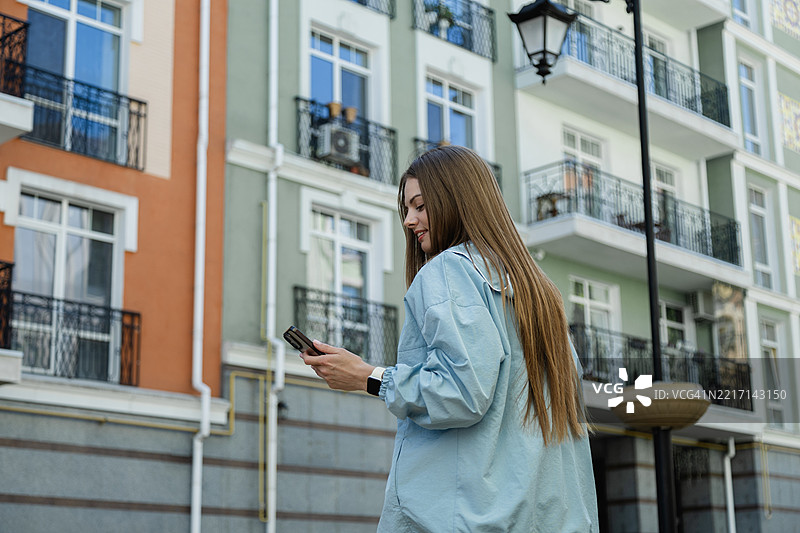 积极的年轻女性在现代住宅楼前使用智能手机。图片素材