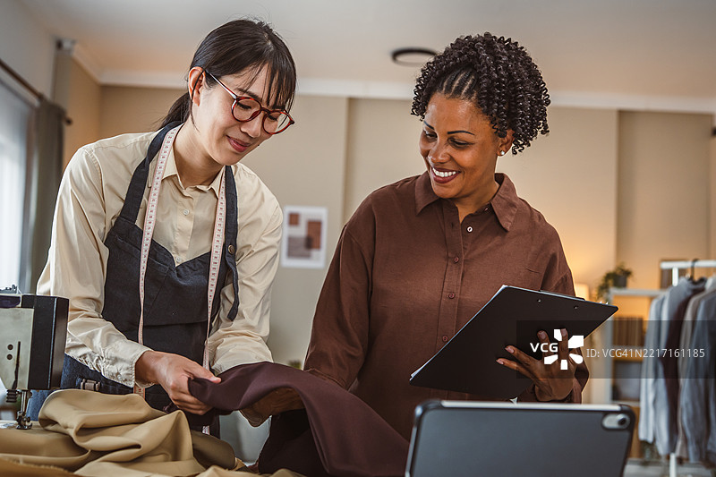 两位女性在一个小型时尚工作室合作——一位脖子上挂着卷尺的日本女性和一位手持夹板的非裔美国经理。图片素材