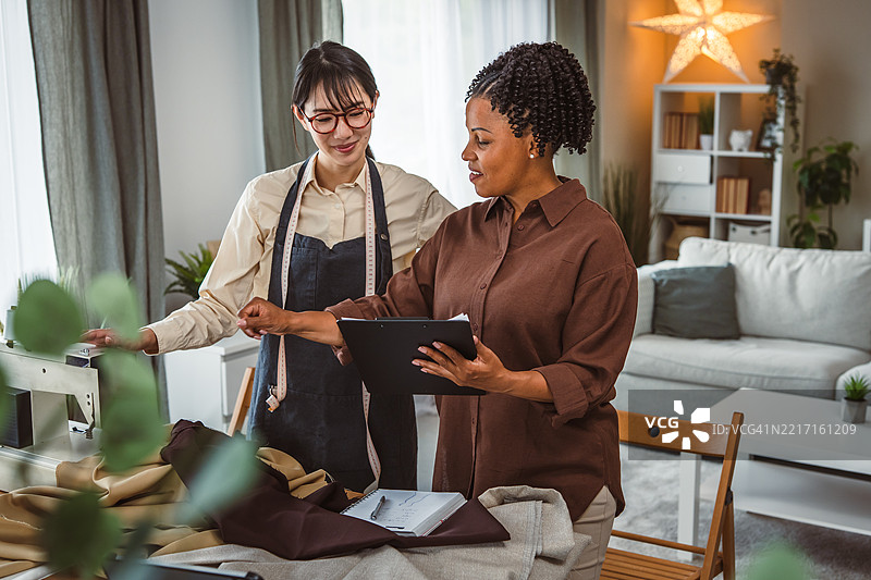 两位女性在一个小型时尚工作室合作——一位脖子上挂着卷尺的日本女性和一位手持夹板的非裔美国经理。图片素材