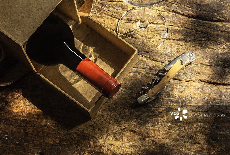 阳光明媚的木质背景上，一瓶红酒从礼品盒中取出，旁边有开瓶器和酒杯。图片素材