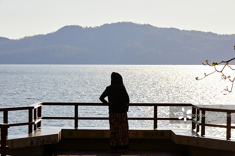 背影剪影的女性站在海边，背景是山脉。图片素材