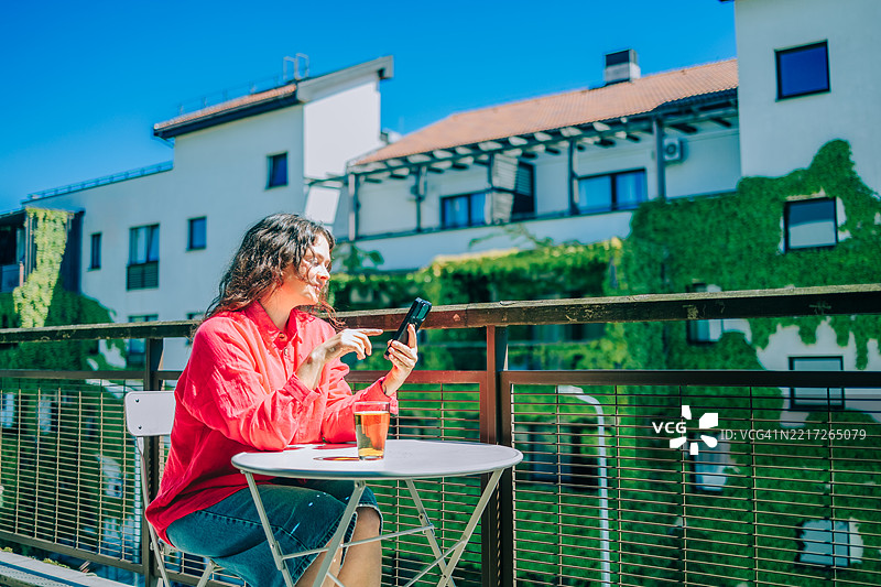 阳光明媚的日子，女人在阳台上放松，手里拿着智能手机和饮料。图片素材
