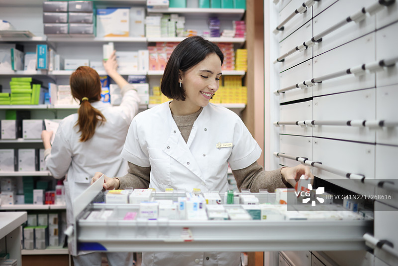 一名女性在药房工作，将药品放入储藏室的抽屉中，背景中有一位同事在工作。图片素材